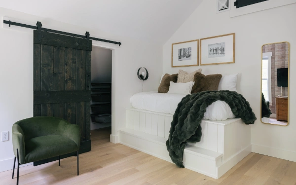 Elevated bed platform with white paneling, dark barn-style sliding door, green accent chair, and layered cozy textiles